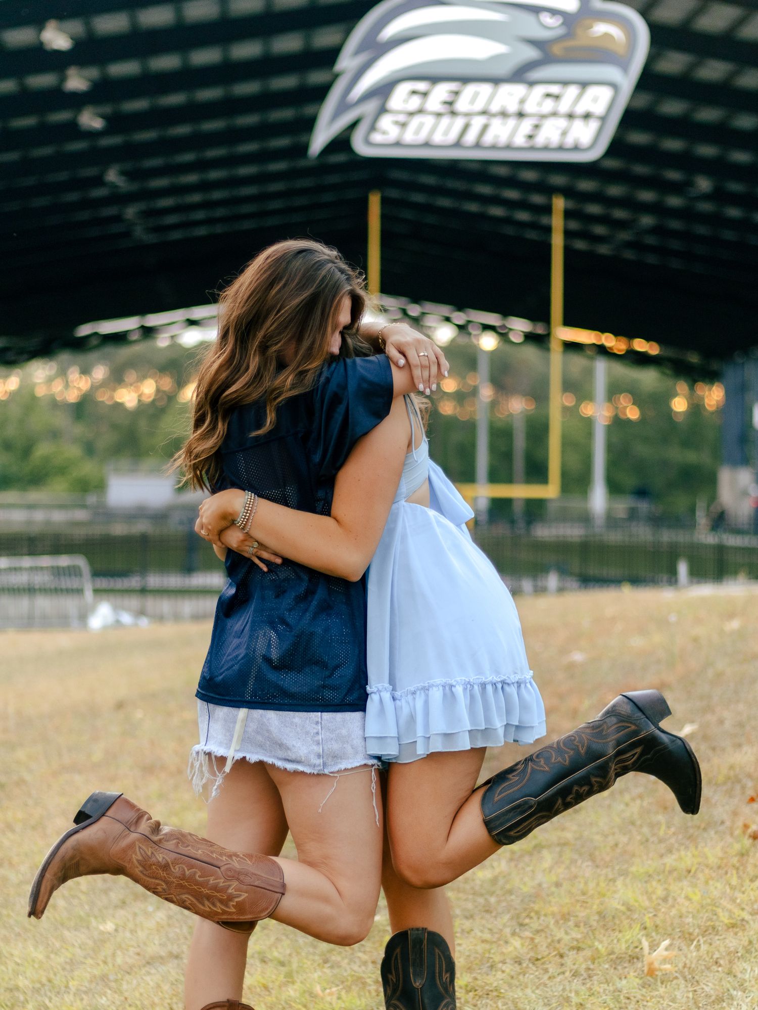 Two women in cowboy boots hug and lift their feet under a Georgia Southern sign at an outdoor venue with string lights.