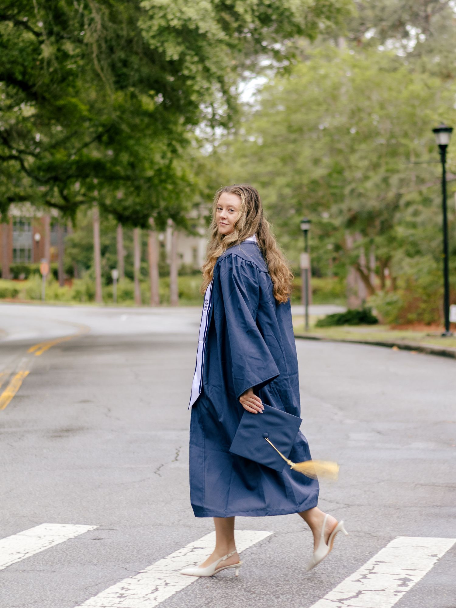 Graduate in a navy cap and gown with a gold tassel crossing a tree-lined campus street at a crosswalk, glancing back over her shoulder.