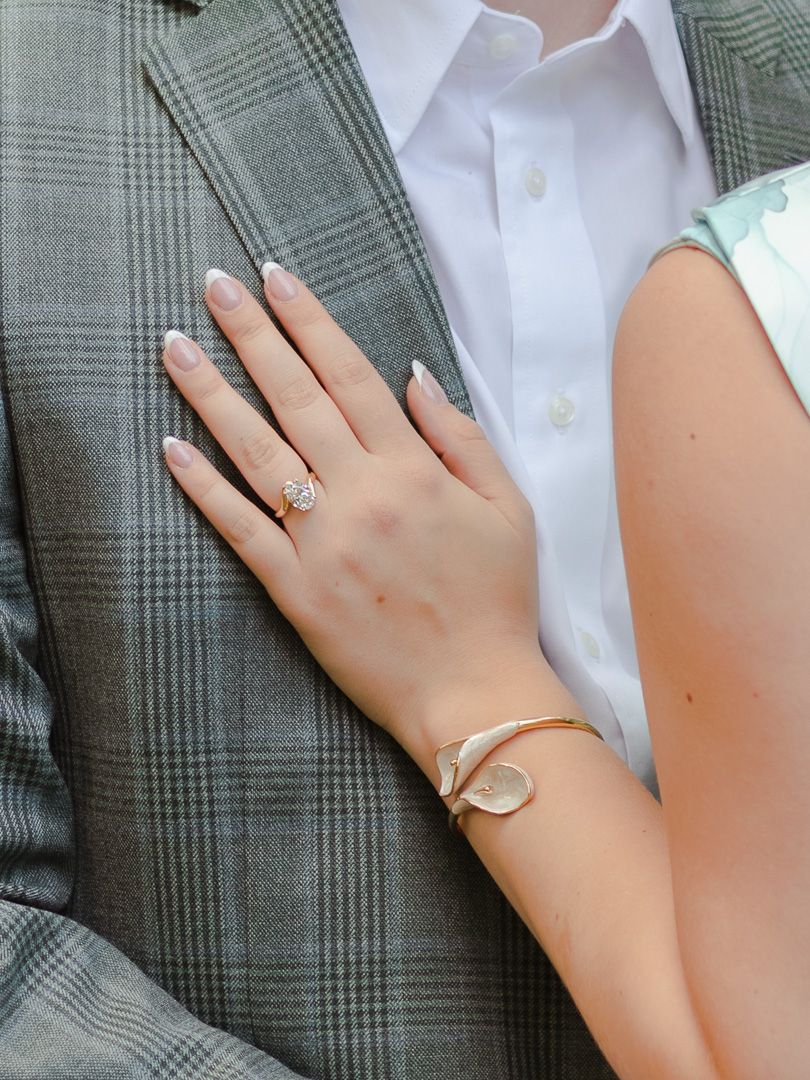 Close-up of a woman's manicured hand with a diamond engagement ring resting on a man's gray plaid blazer and white dress shirt.