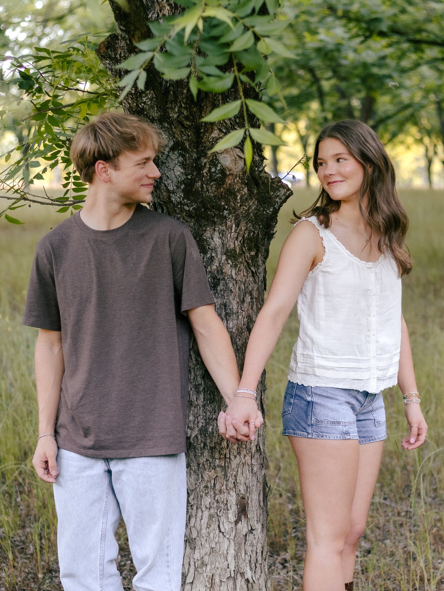 Young couple holding hands and smiling beside a large tree trunk in a sunny grassy field, wearing casual summer outfits.