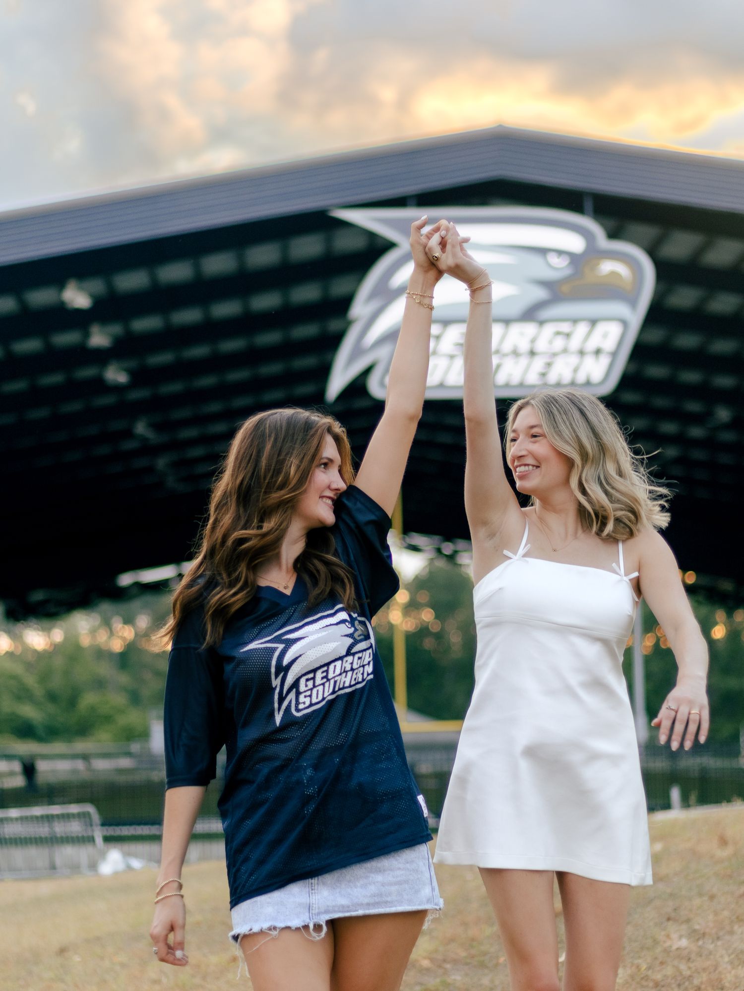 Two women in Georgia Southern gear hold hands raised in front of the Georgia Southern Eagles sign at sunset.