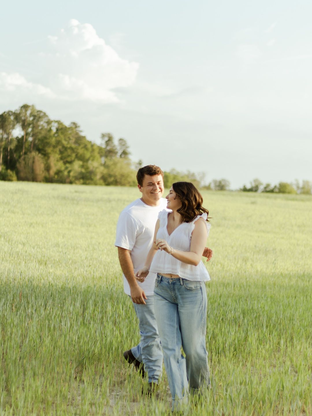 Smiling couple in white tops and jeans walk arm in arm through a sunlit green grass field with trees under a soft blue sky.