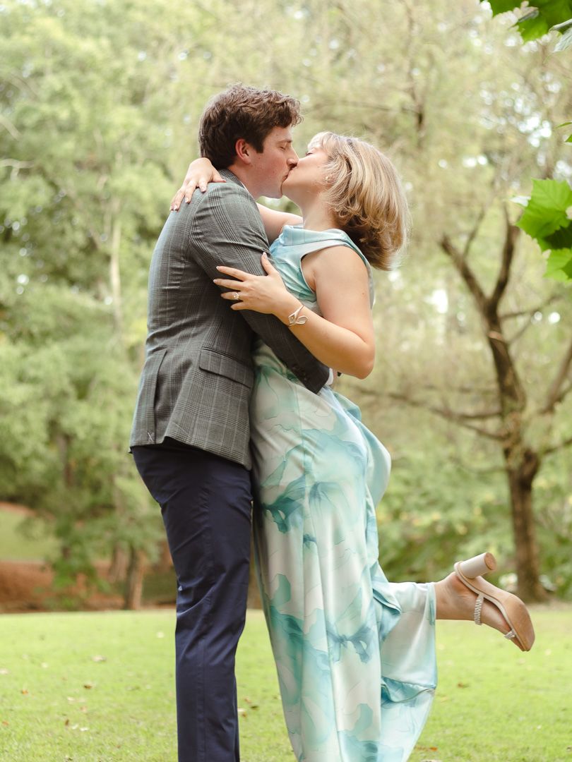 A couple kisses on a lush green lawn as the woman in a watercolor-print dress playfully lifts her heel behind her.
