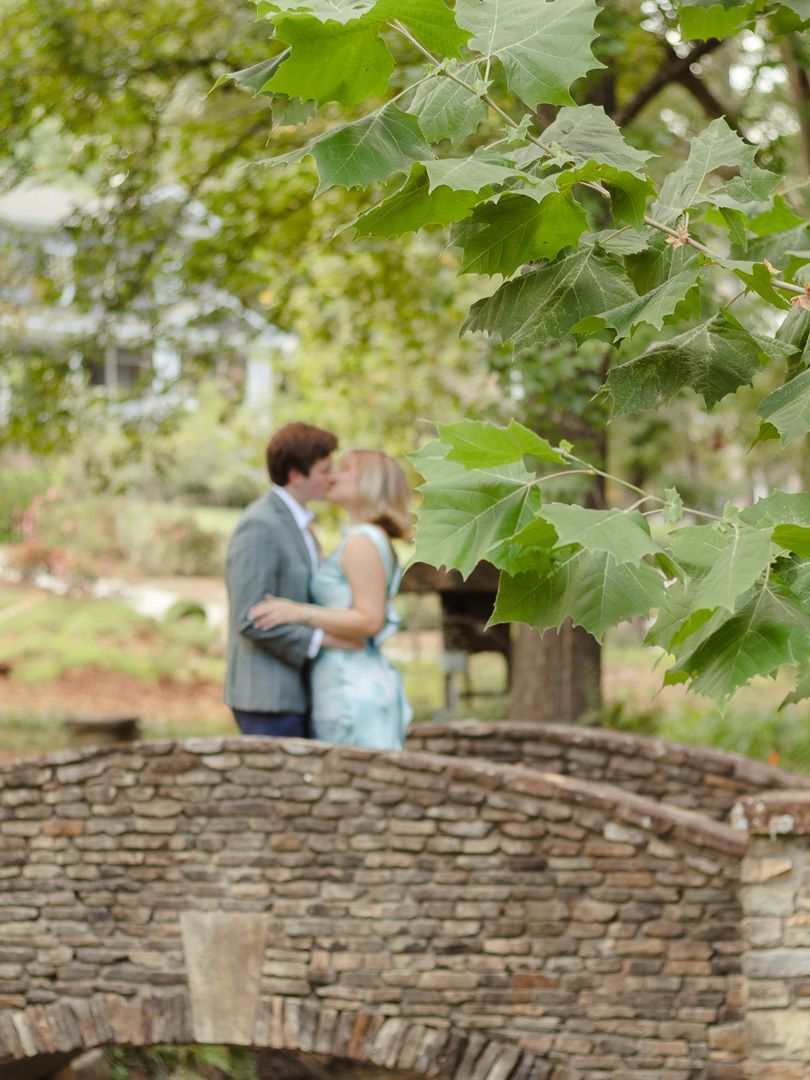 Couple sharing a kiss near a stone arched bridge in a sunlit park, framed by green leaves in an engagement-style photo.