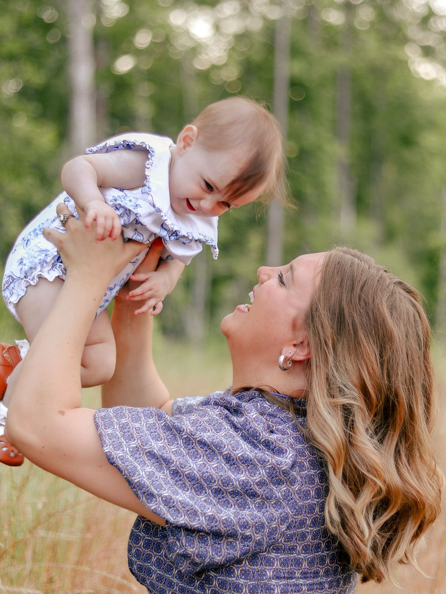Smiling woman with long wavy hair lifts a laughing baby in a floral dress into the air in a sunny grassy field with trees.