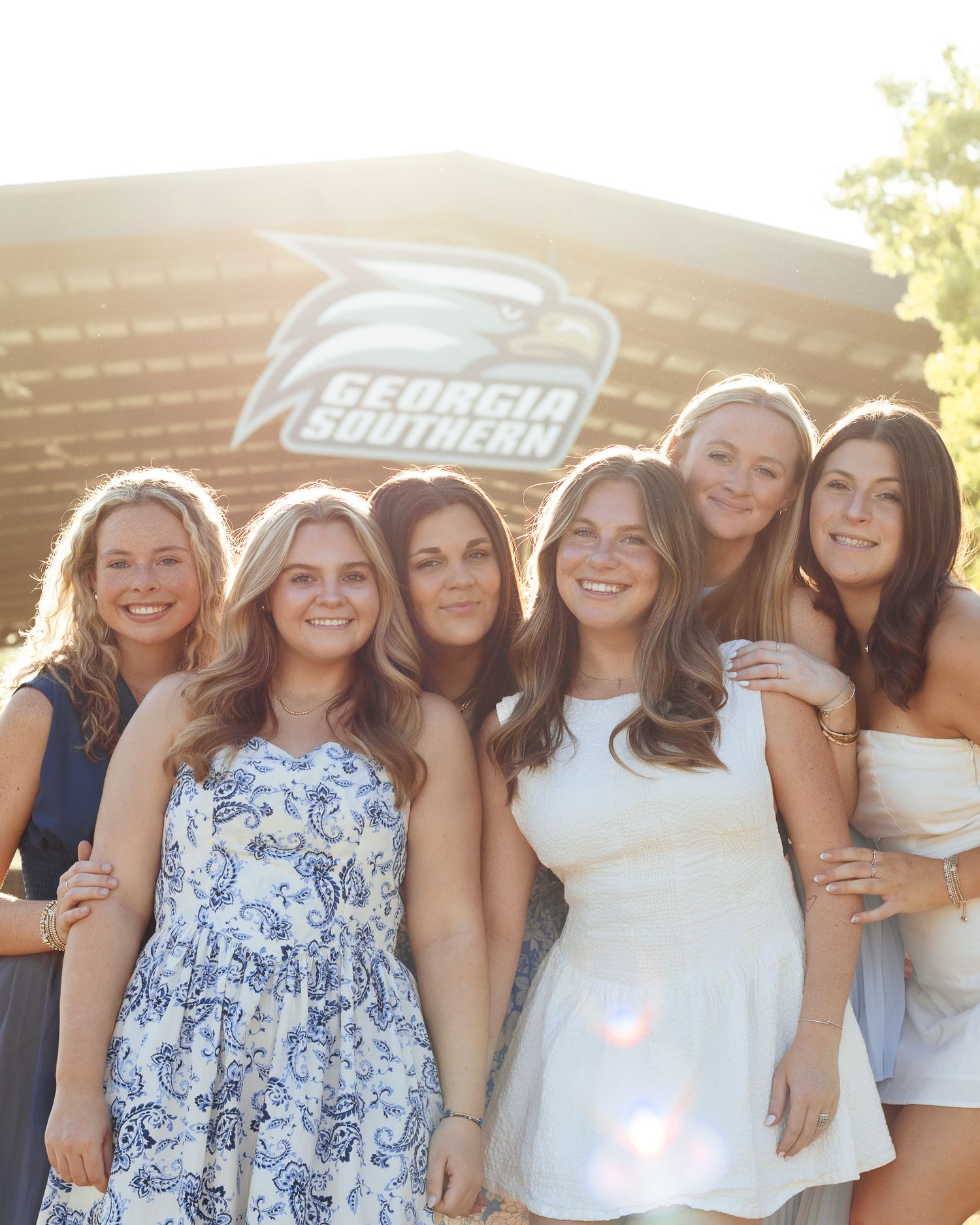 Six smiling young women in summer dresses pose together outdoors in warm sunlight in front of a Georgia Southern University building.