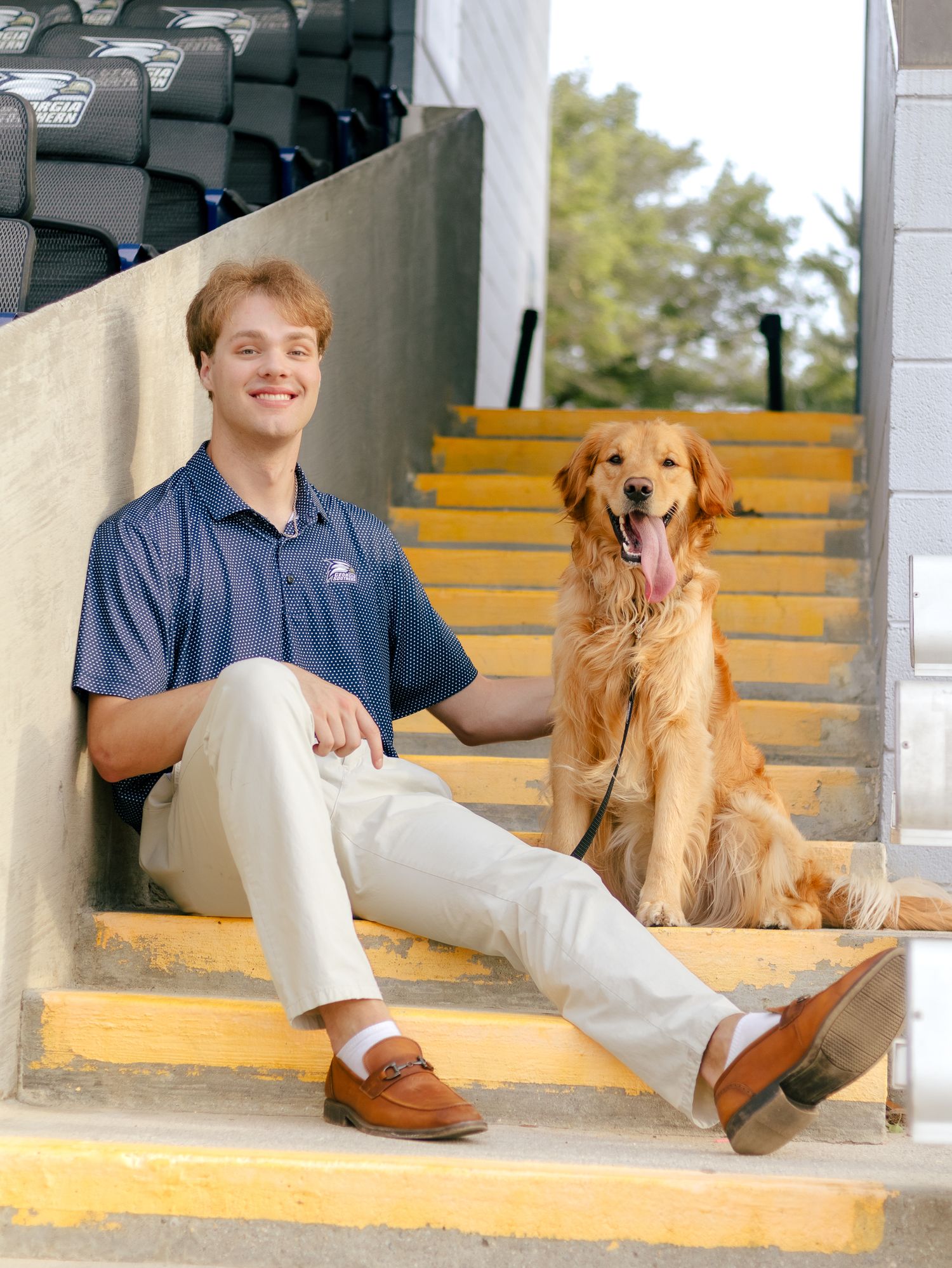 Smiling young man in a navy polo and khakis sits on yellow stadium steps beside a happy golden retriever with its tongue out.