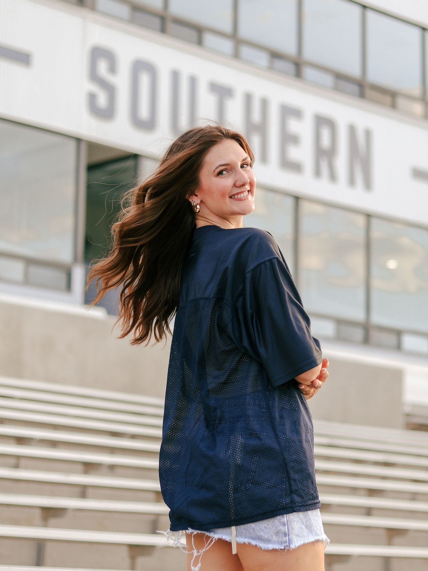 Smiling young woman with long brown hair wearing a navy jersey and denim shorts poses in front of stadium bleachers and a Southern sign.