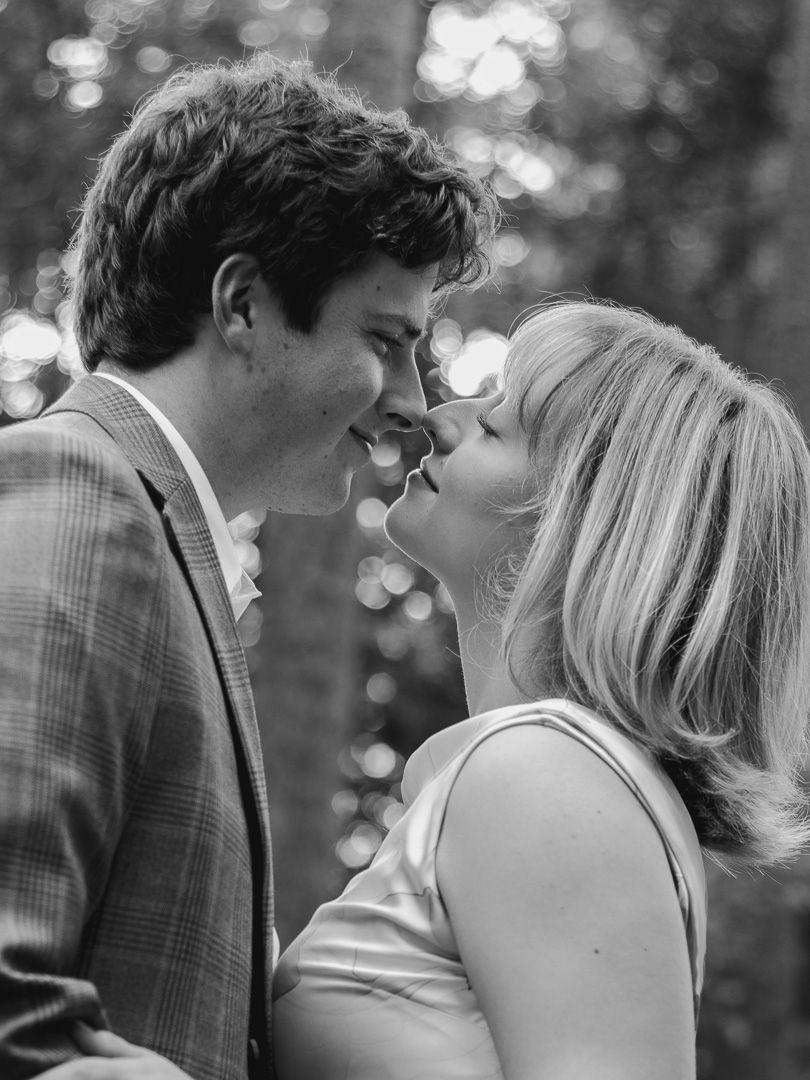 Black and white portrait of a smiling couple about to kiss outdoors, with soft blurred trees glowing in the background.