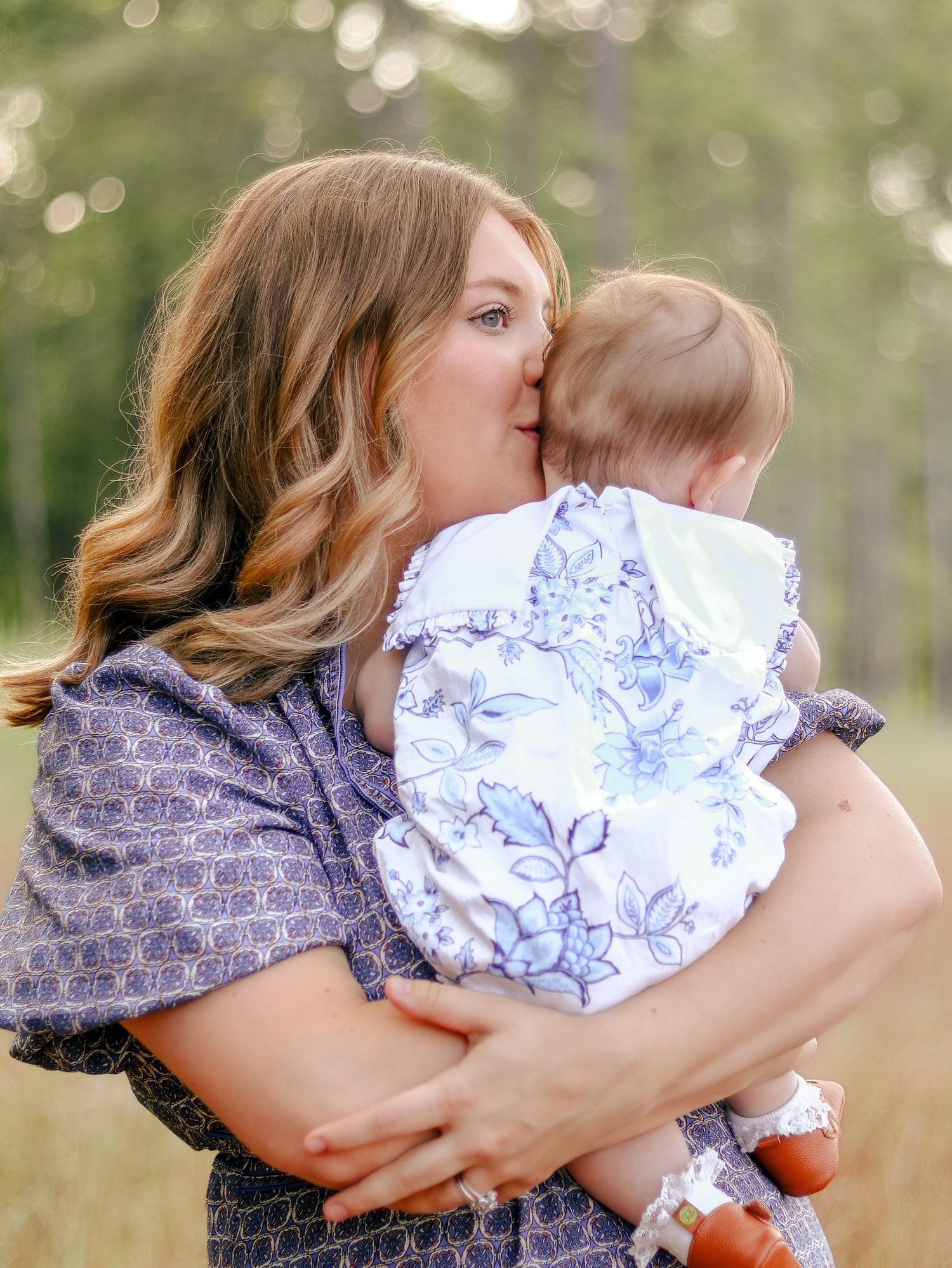 A woman with wavy hair gently kisses a baby in a blue floral outfit while holding her close in a sunlit grassy field with trees.