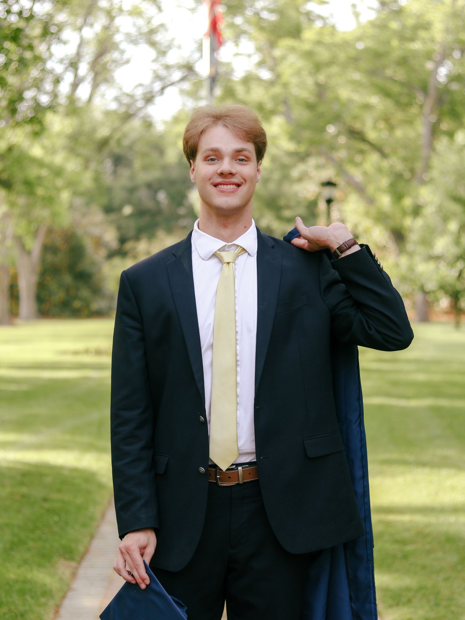 Smiling young man in a dark suit and yellow tie holds a navy graduation gown over his shoulder on a sunny, tree-lined lawn.
