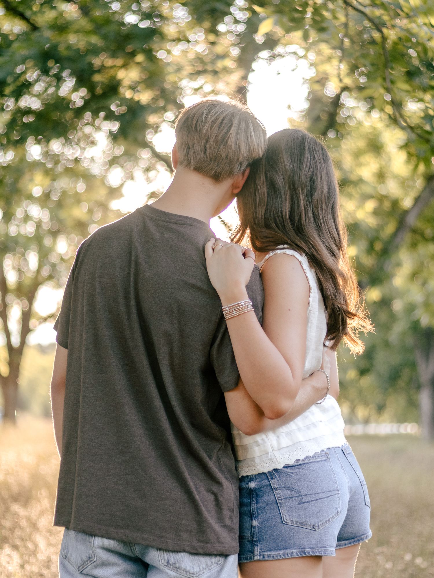Couple embracing with their backs to the camera, standing in a sunlit park surrounded by trees at golden hour.