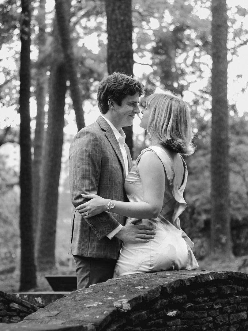 Black and white photo of a couple embracing and about to kiss on a stone bridge surrounded by tall trees.
