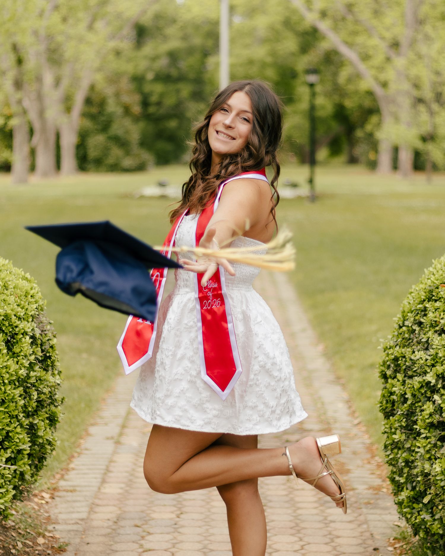 Smiling graduate in a white dress and red sash poses on a brick path, holding a navy cap and tassel while kicking up one heel.