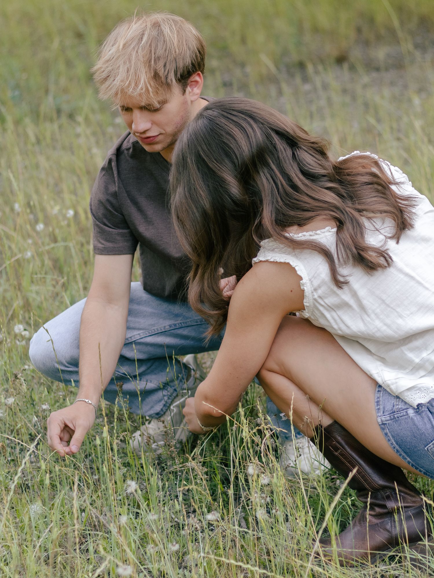 Young man and woman crouching together in a sunny meadow of tall grass and wildflowers, looking closely at the ground.