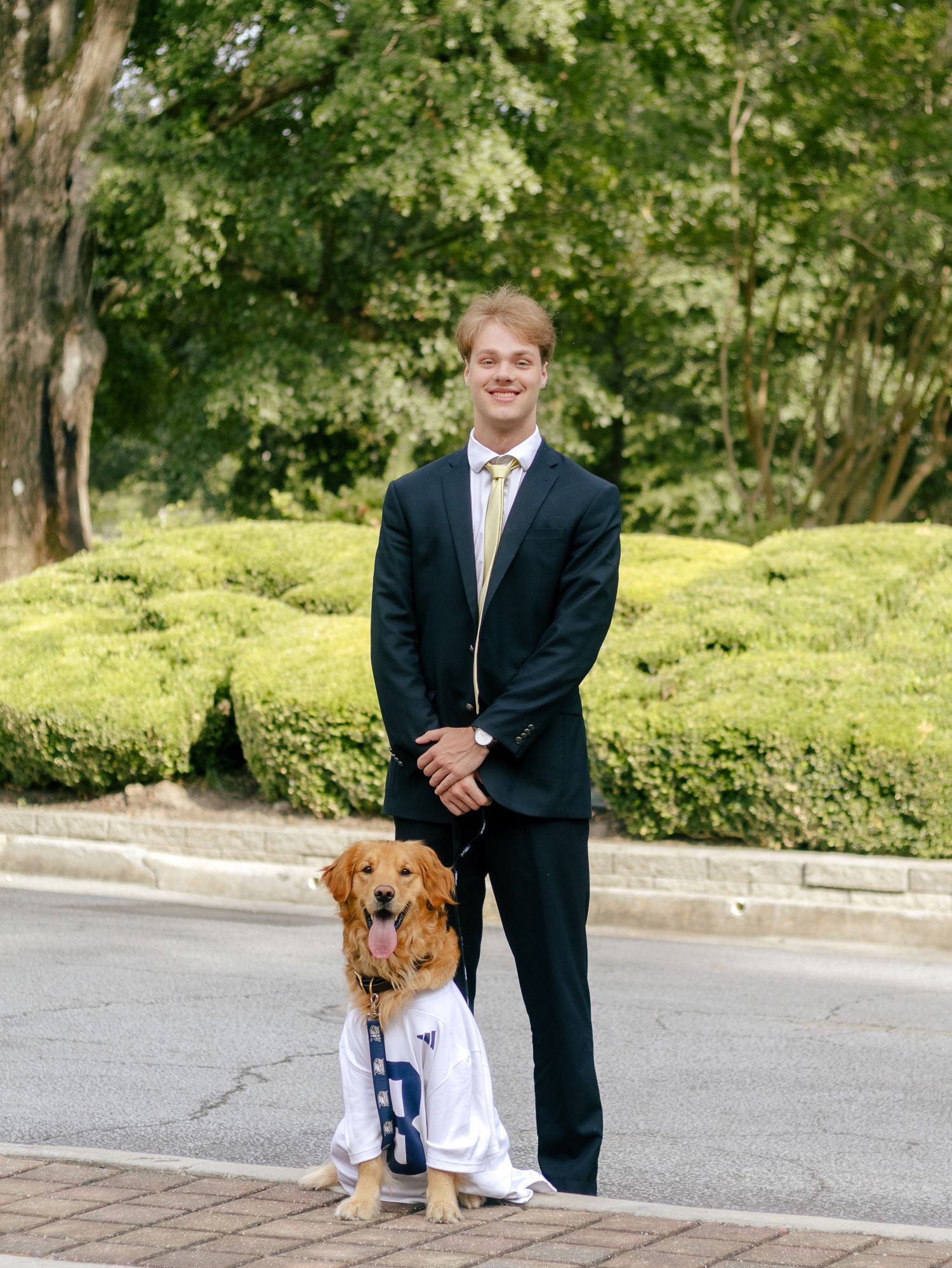 Young man in a dark suit and tie stands on a brick sidewalk beside a golden retriever wearing a white jersey, with trimmed hedges behind.