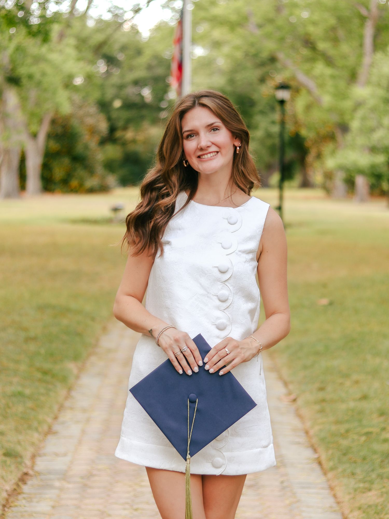 Smiling young woman in a white sleeveless button dress holds a navy graduation cap while standing on a brick path in a sunny park.