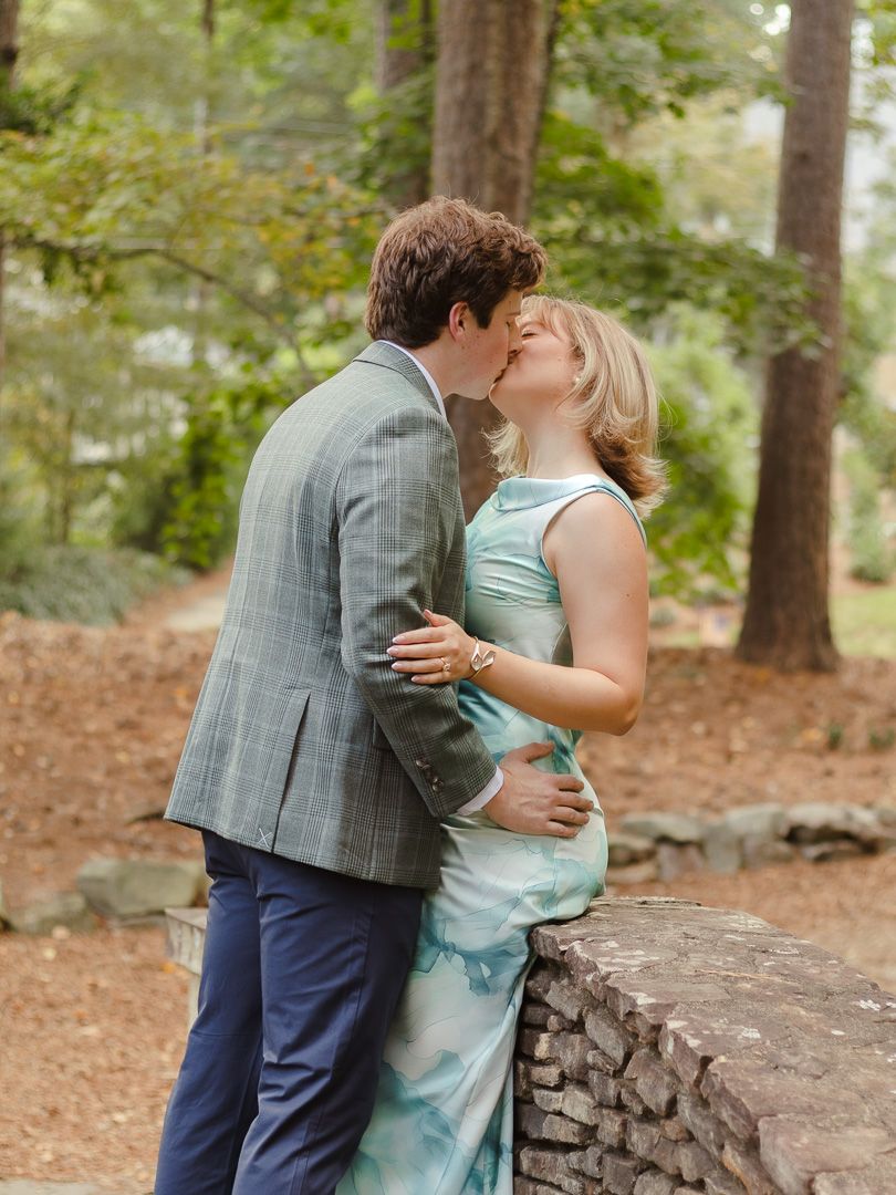 A couple shares a romantic kiss beside a stone wall in a sunlit wooded park, the man in a plaid blazer and the woman in a teal dress.