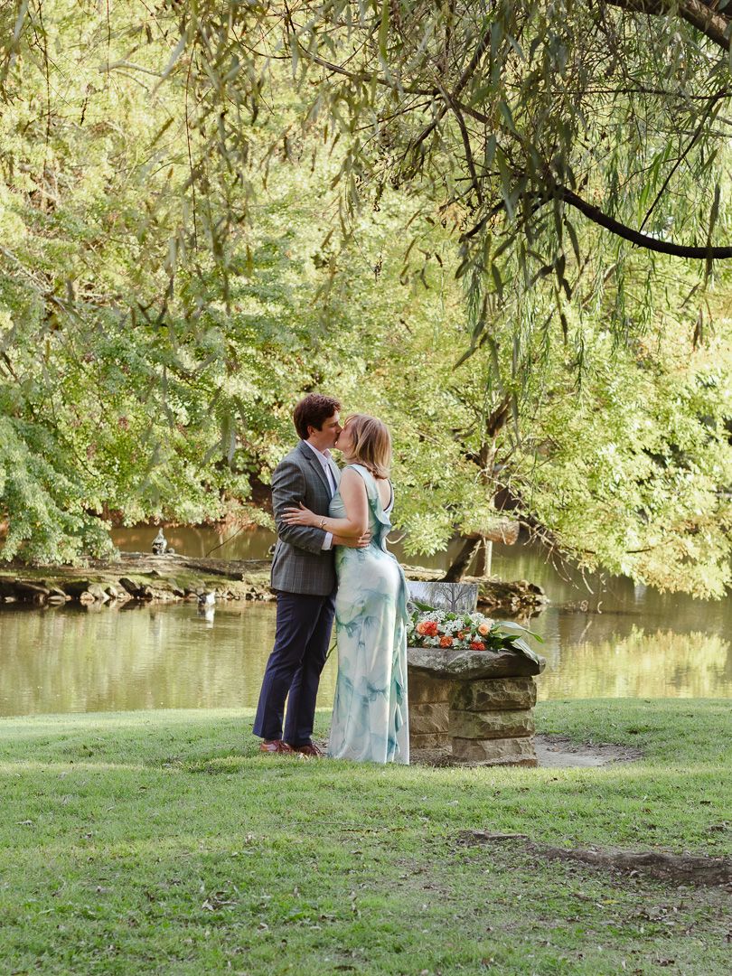 Couple in a romantic embrace under a willow tree beside a calm pond, standing on green grass near a stone ledge with flowers.