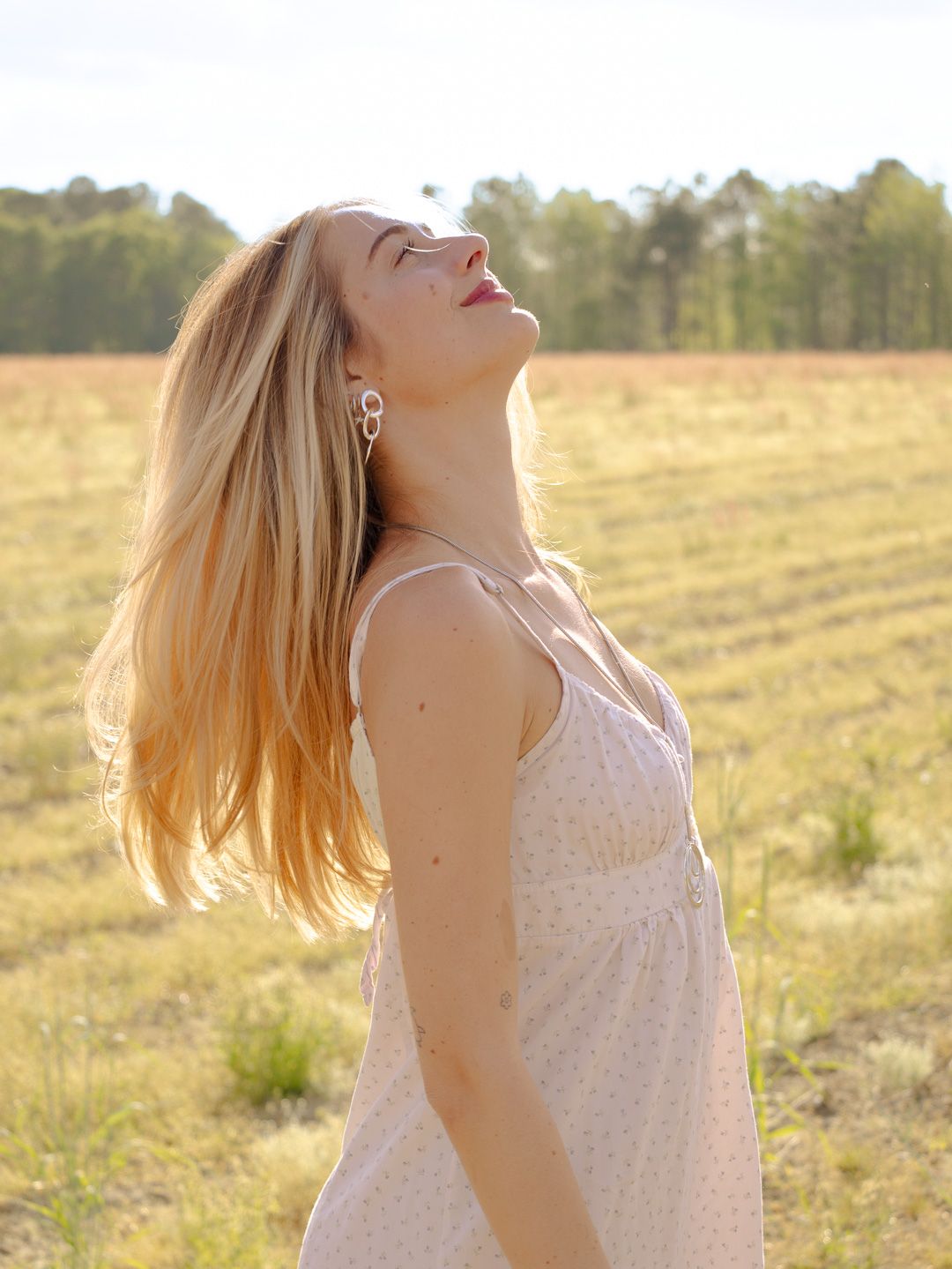 Woman with long blonde hair tilts her head back in golden sunlight, wearing a pale floral sundress in a grassy field with trees behind.