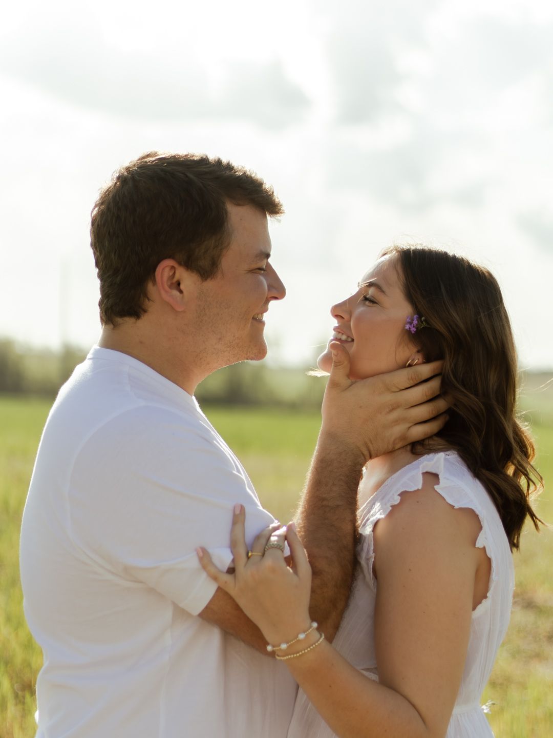 Smiling couple in white outfits embrace face to face in a sunny green field, perfect for engagement or romantic couple photography.