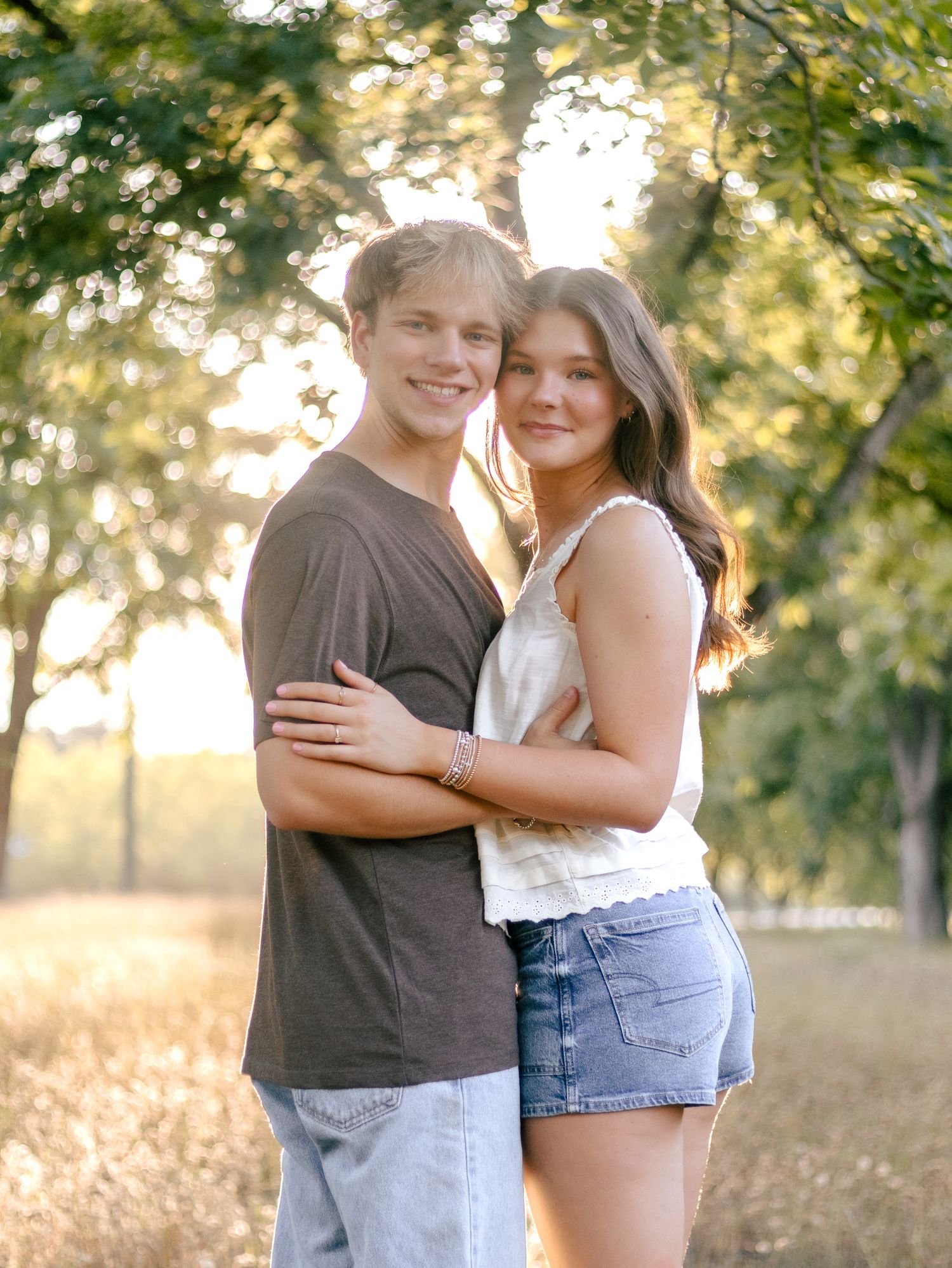 Smiling young couple embracing outdoors in a sunlit field with trees, he wears a brown tee and jeans, she a white top and denim shorts.