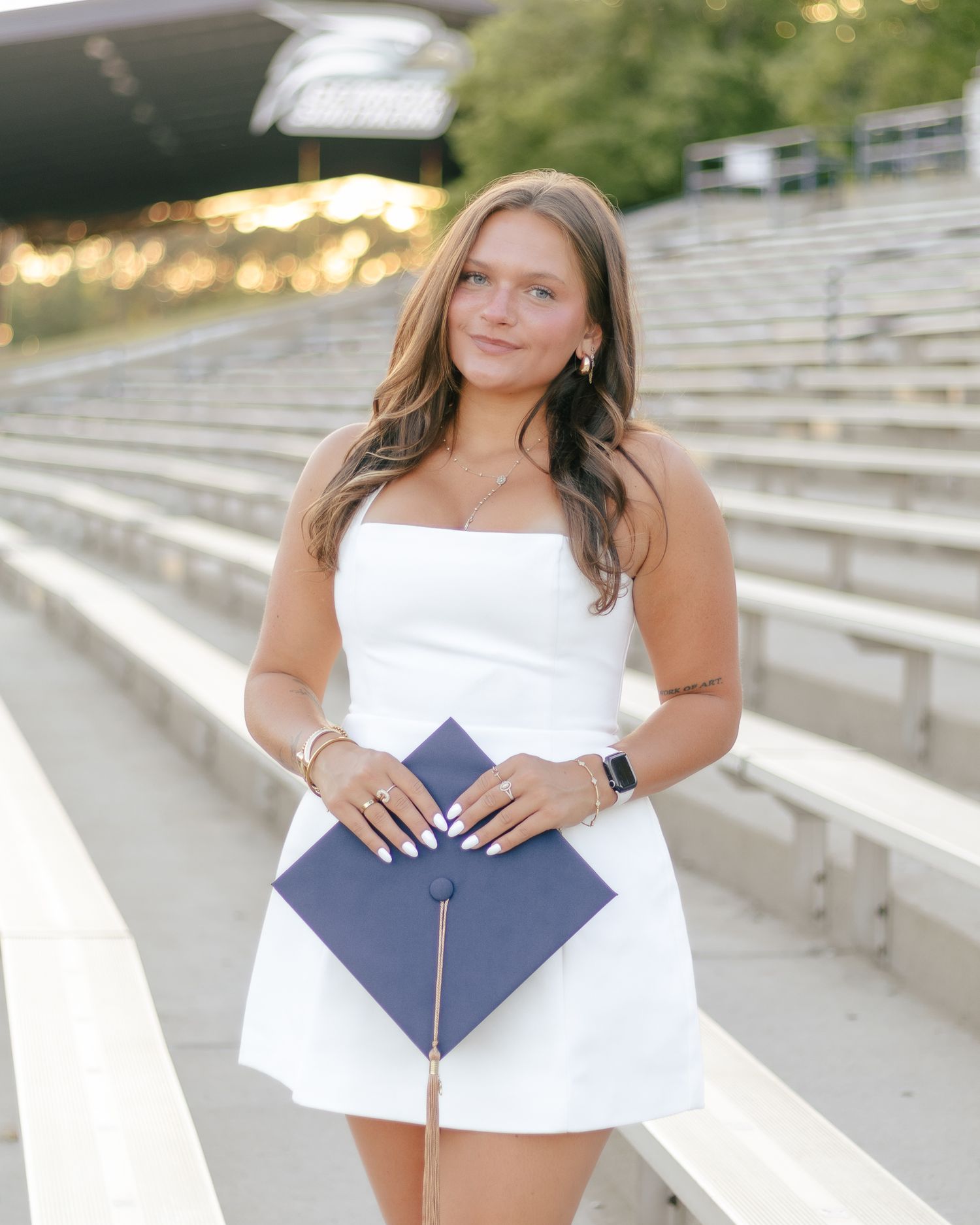 Smiling graduate in a white dress holds a navy graduation cap with a tassel while standing on empty stadium bleachers.