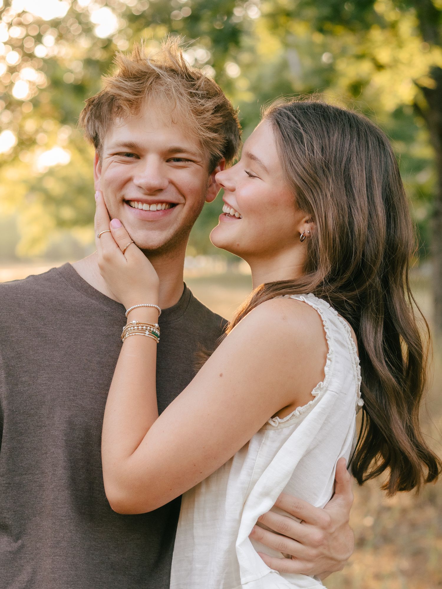 Smiling young couple embracing outdoors as a woman gently touches a man's cheek, with sunlit green trees softly blurred behind them.