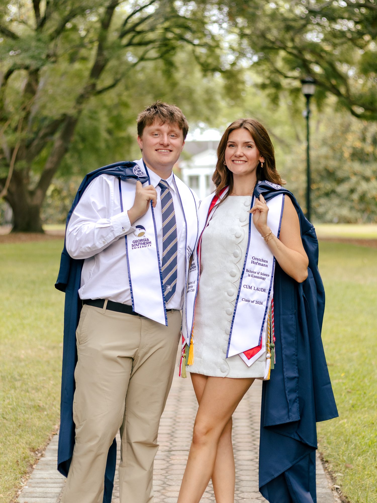 Two smiling graduates in navy gowns draped over their shoulders and white honor stoles pose on a brick path beneath tall oak trees.