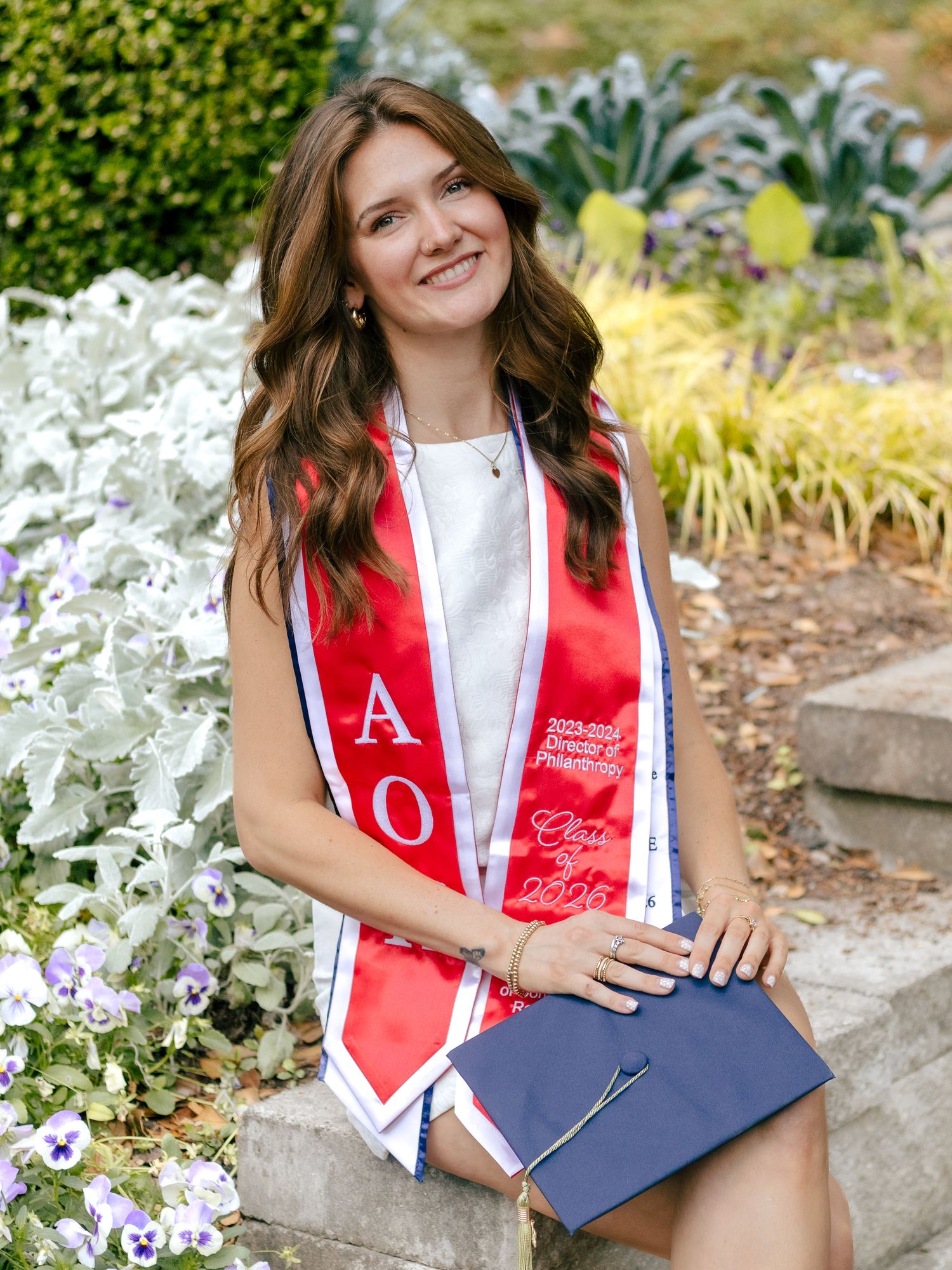 Smiling graduate in a white dress and red sorority graduation stole sits on stone steps holding a navy cap beside purple pansies.