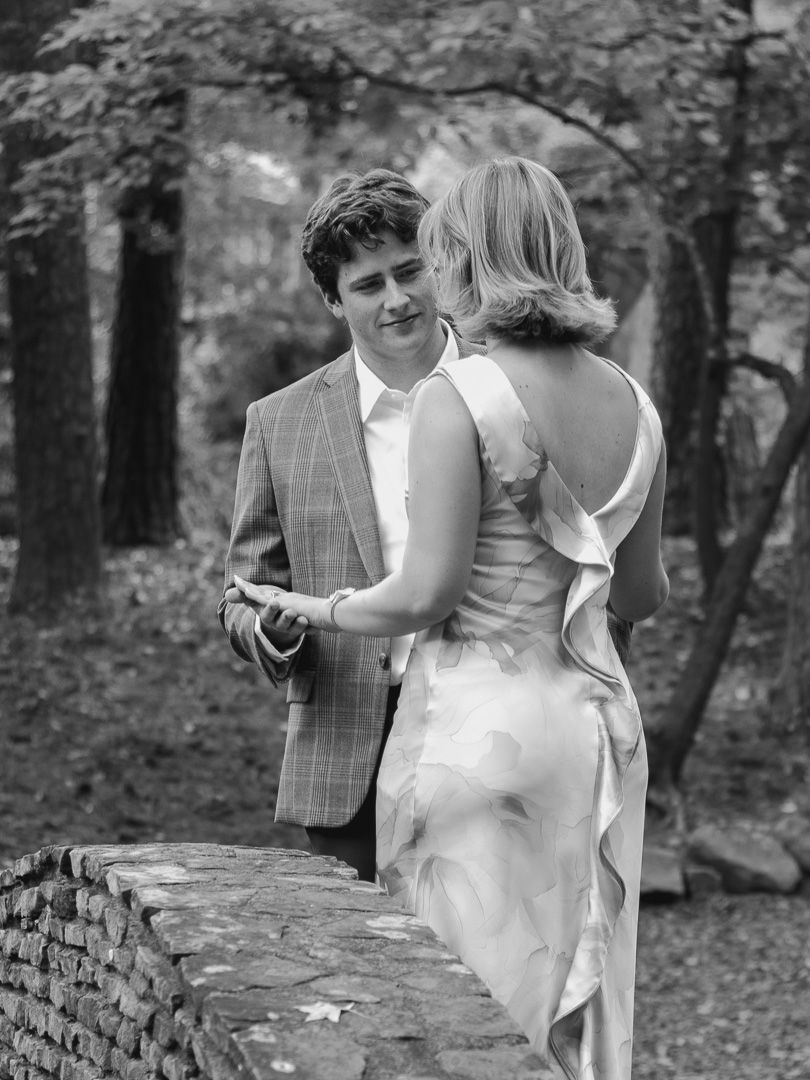 Black and white portrait of a couple dancing close together on a stone bridge in a wooded park, he in a plaid blazer, she in a gown.