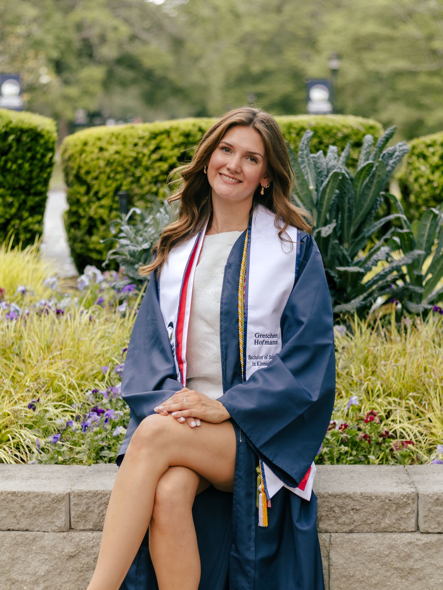 Smiling graduate in a navy cap and gown with a white honor stole sits on a stone ledge in front of a landscaped garden.