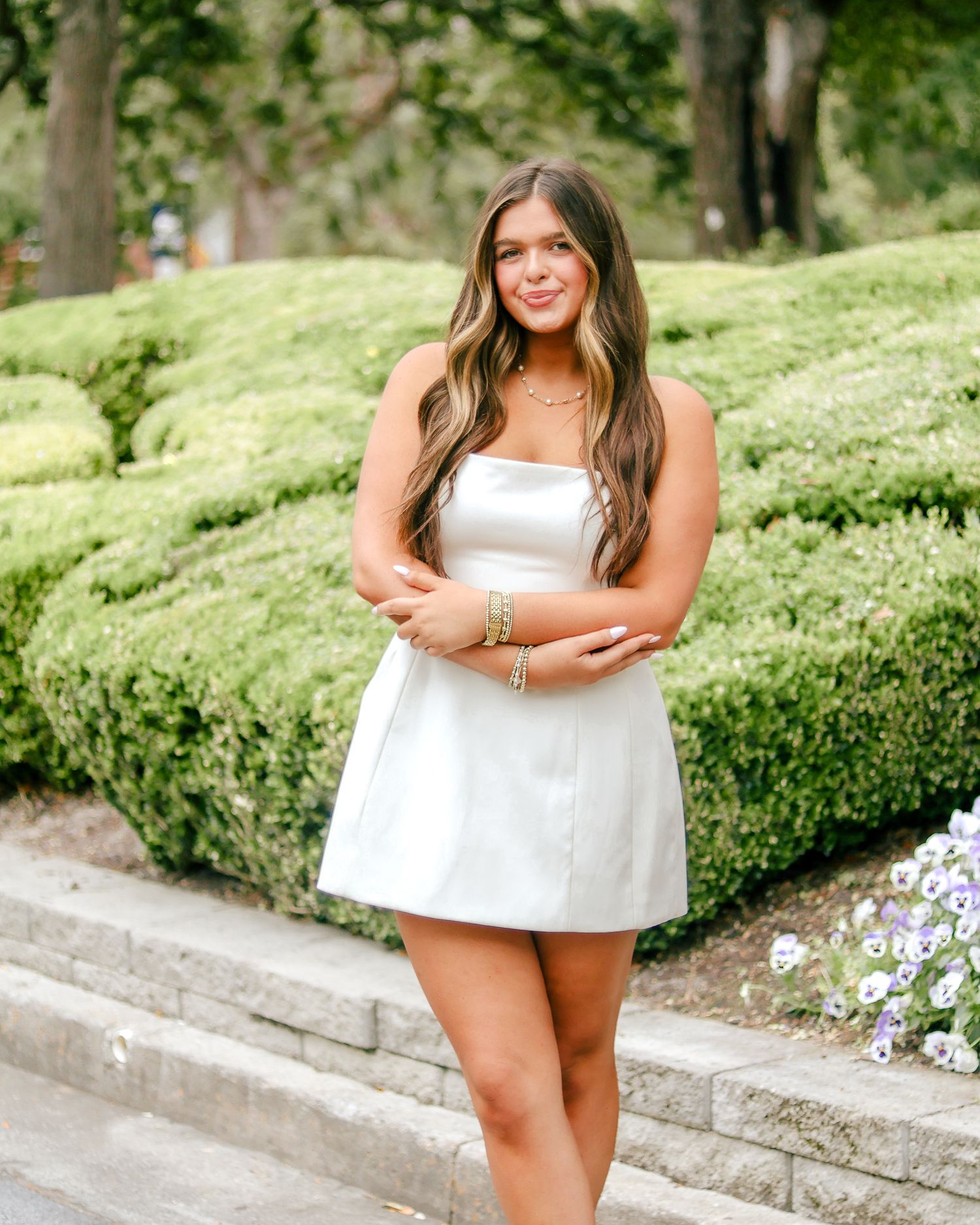 Young woman with long wavy hair poses in a white satin mini dress and gold bracelets beside trimmed green hedges and white flowers.