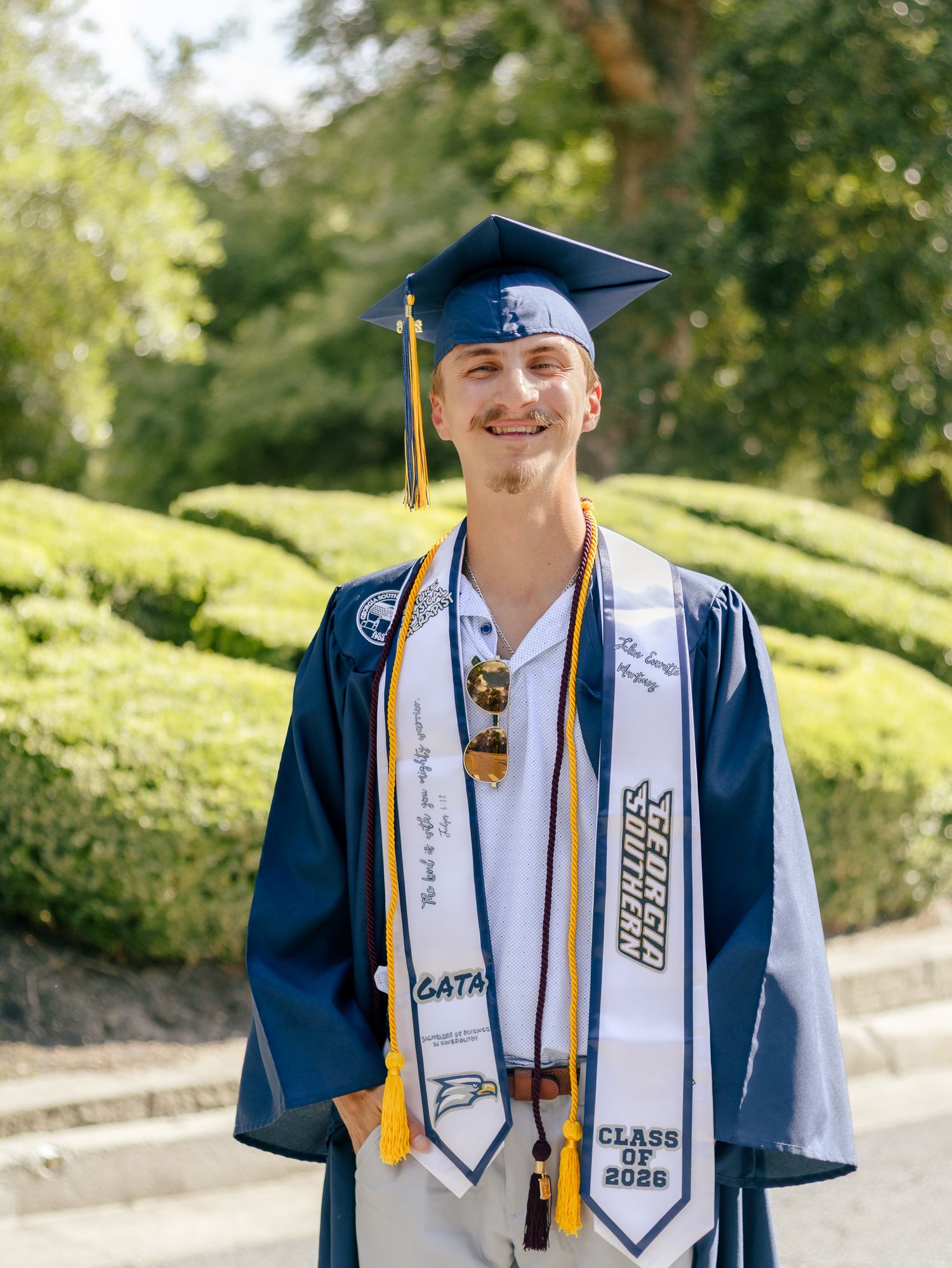 Smiling graduate in a navy cap and gown with gold tassel and white Class of 2022 stole stands outdoors in front of trimmed green hedges.