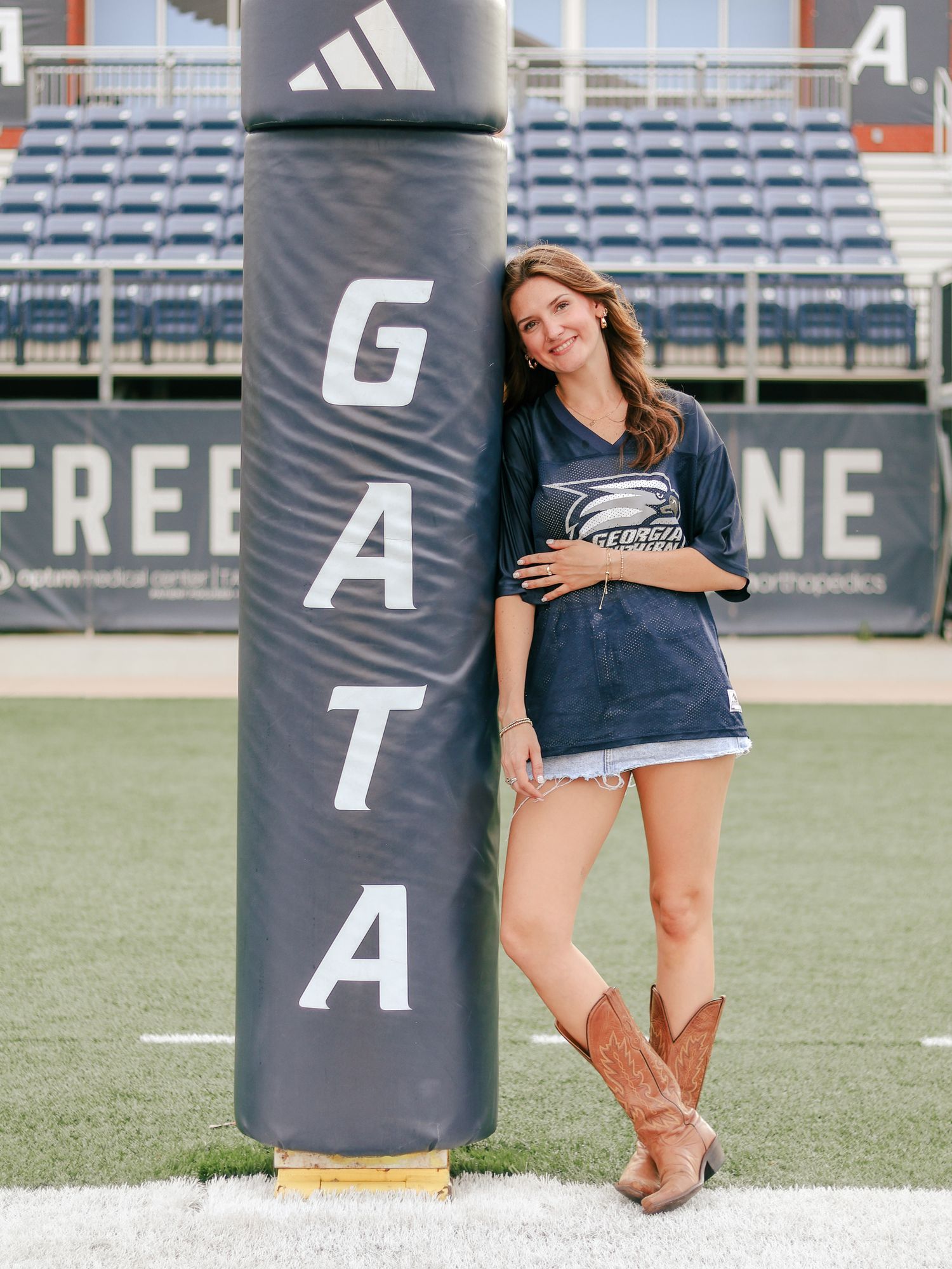 Smiling woman in a navy graphic tee, denim shorts and cowboy boots leans against a padded GATA football goalpost on a stadium field.