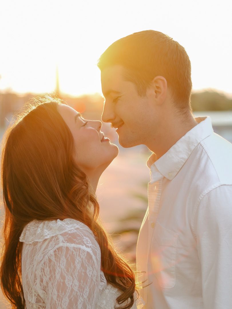 Smiling couple standing nose to nose in warm golden sunset light, she wears a white lace top and he a white button-up shirt.
