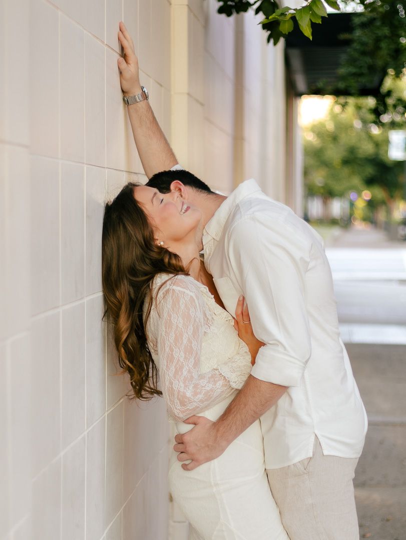 Romantic couple in white outfits embracing against a light stone wall as the man leans in, with a tree-lined street behind them.