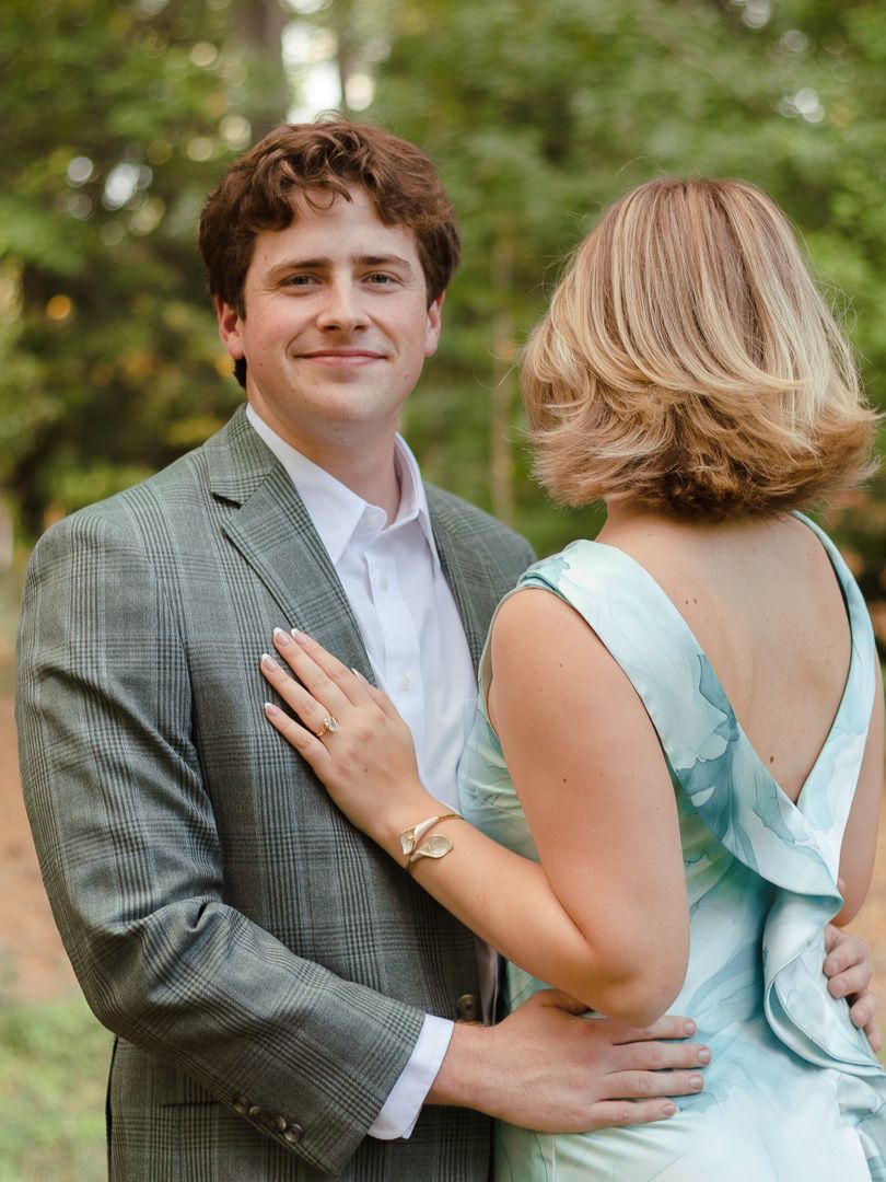 Smiling man in a gray plaid blazer embraces a woman with a bob haircut in a light blue dress outdoors among green trees.