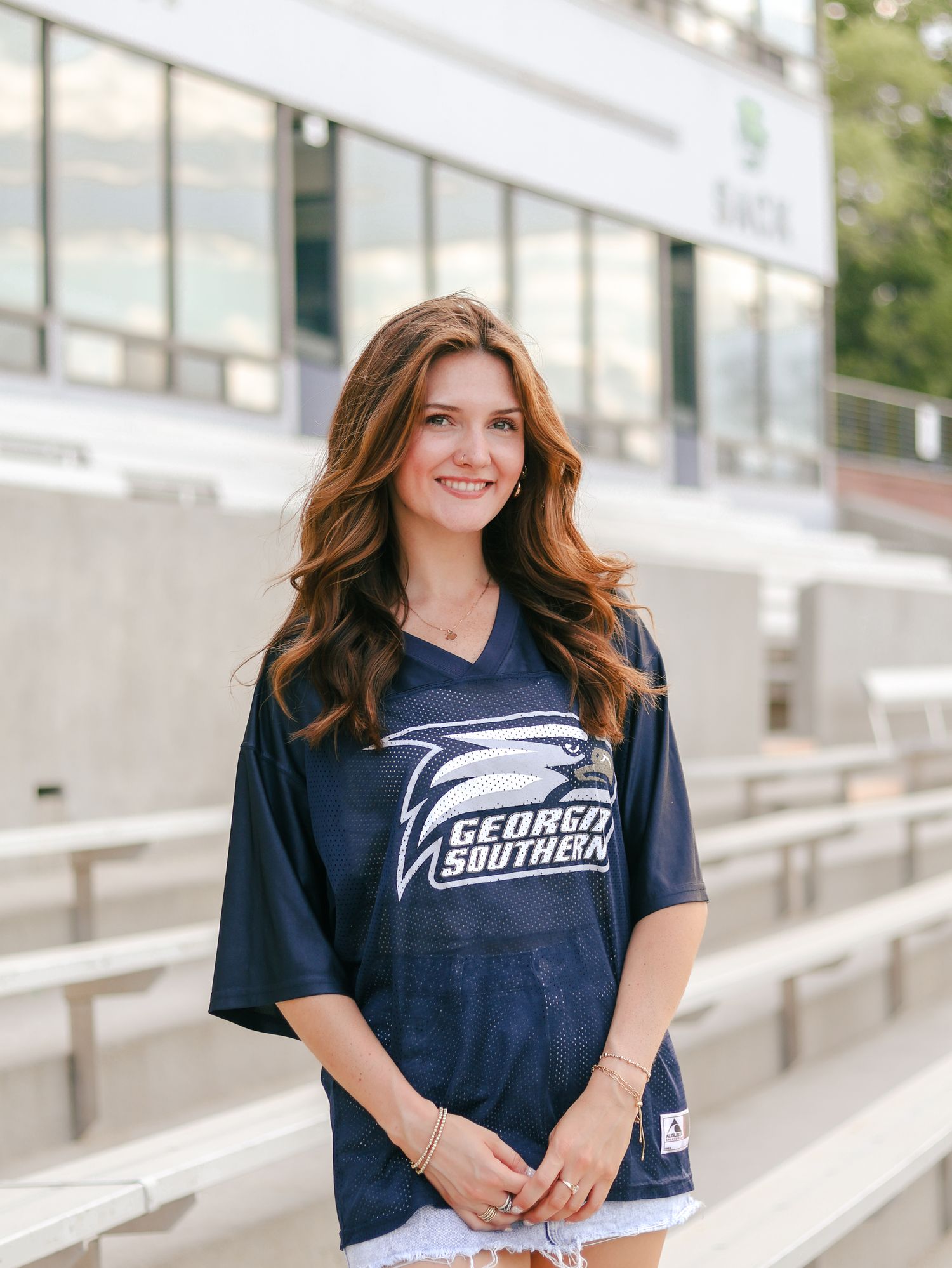 Smiling woman with long wavy auburn hair wearing a navy Georgia Southern jersey, standing in front of empty stadium bleachers.