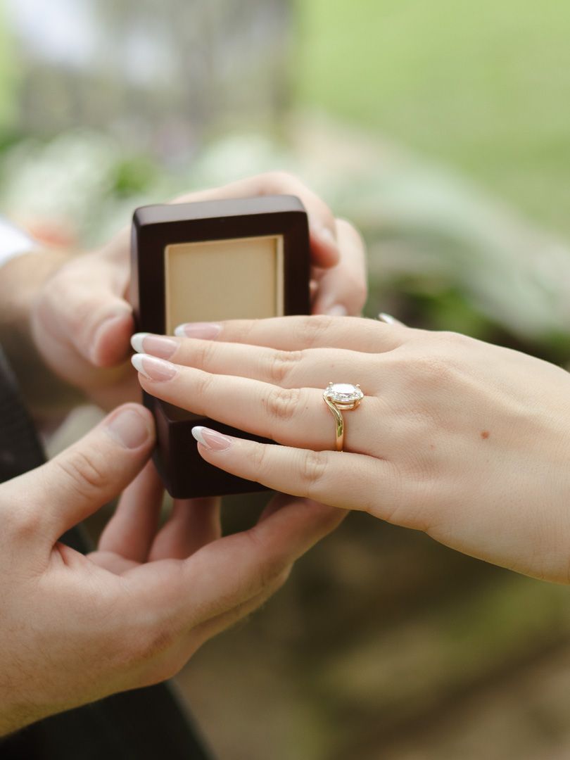 A man presents an open ring box as a woman's hand, wearing a sparkling oval diamond engagement ring, reaches toward it outdoors.