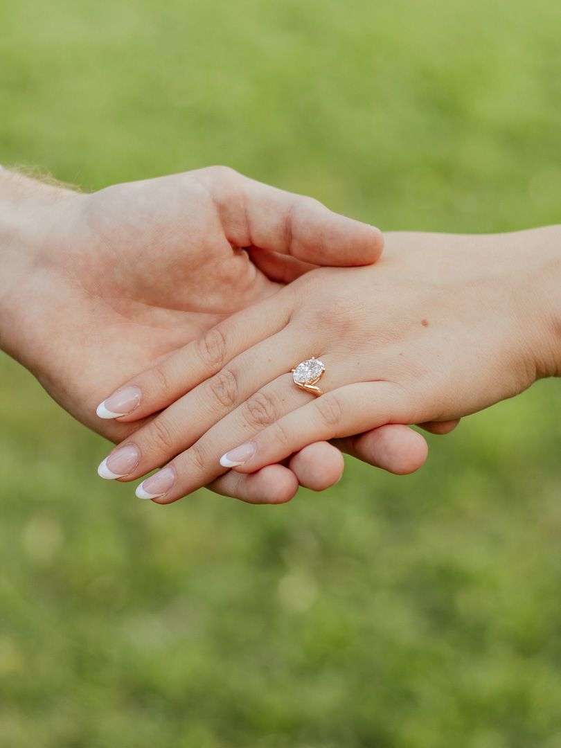 Close-up of a man's hand holding a woman's hand wearing a sparkling engagement ring against a green grass background.