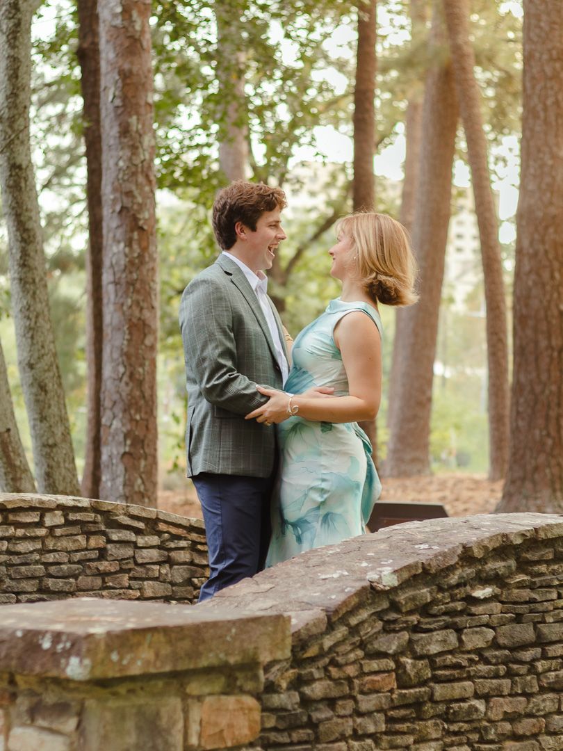Smiling couple in a gray blazer and teal dress embrace beside a curved stone wall in a sunlit wooded park.