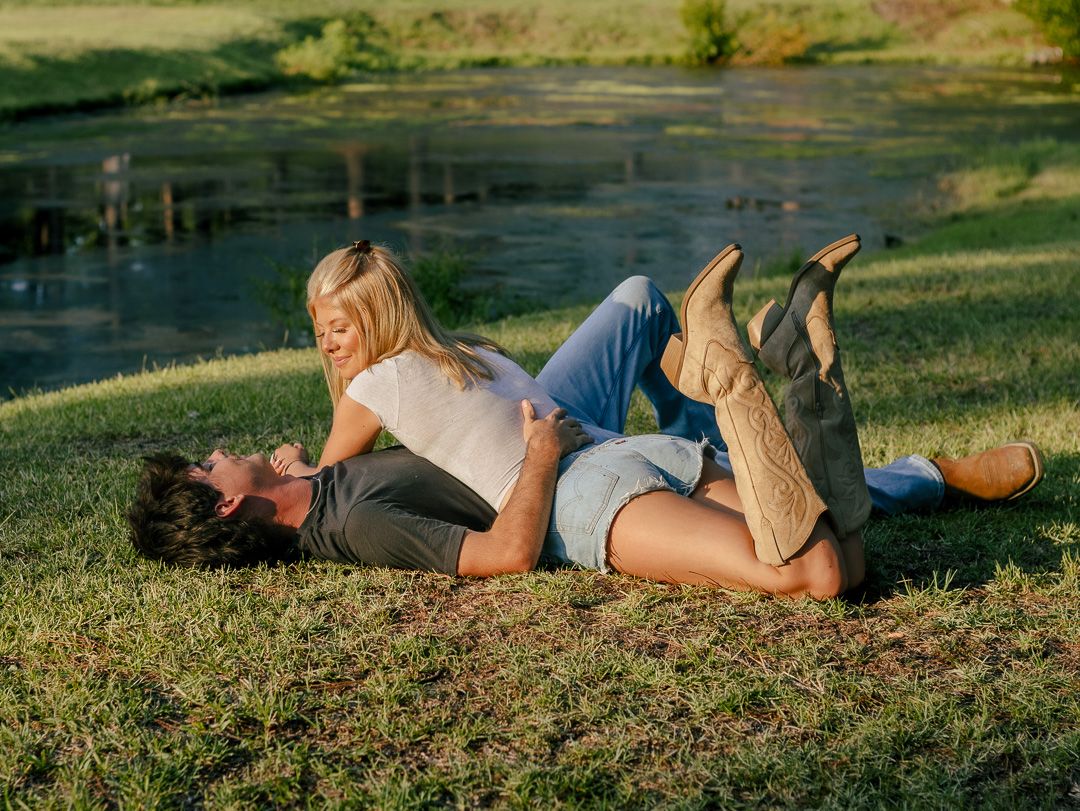 A couple lies on the grass beside a calm pond, the woman in a white top and cowboy boots resting against the man's side.