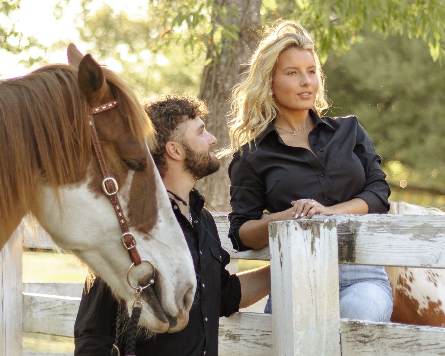 A blonde woman and a bearded man in black shirts lean on a white wooden fence beside a brown and white horse in a sunny pasture.