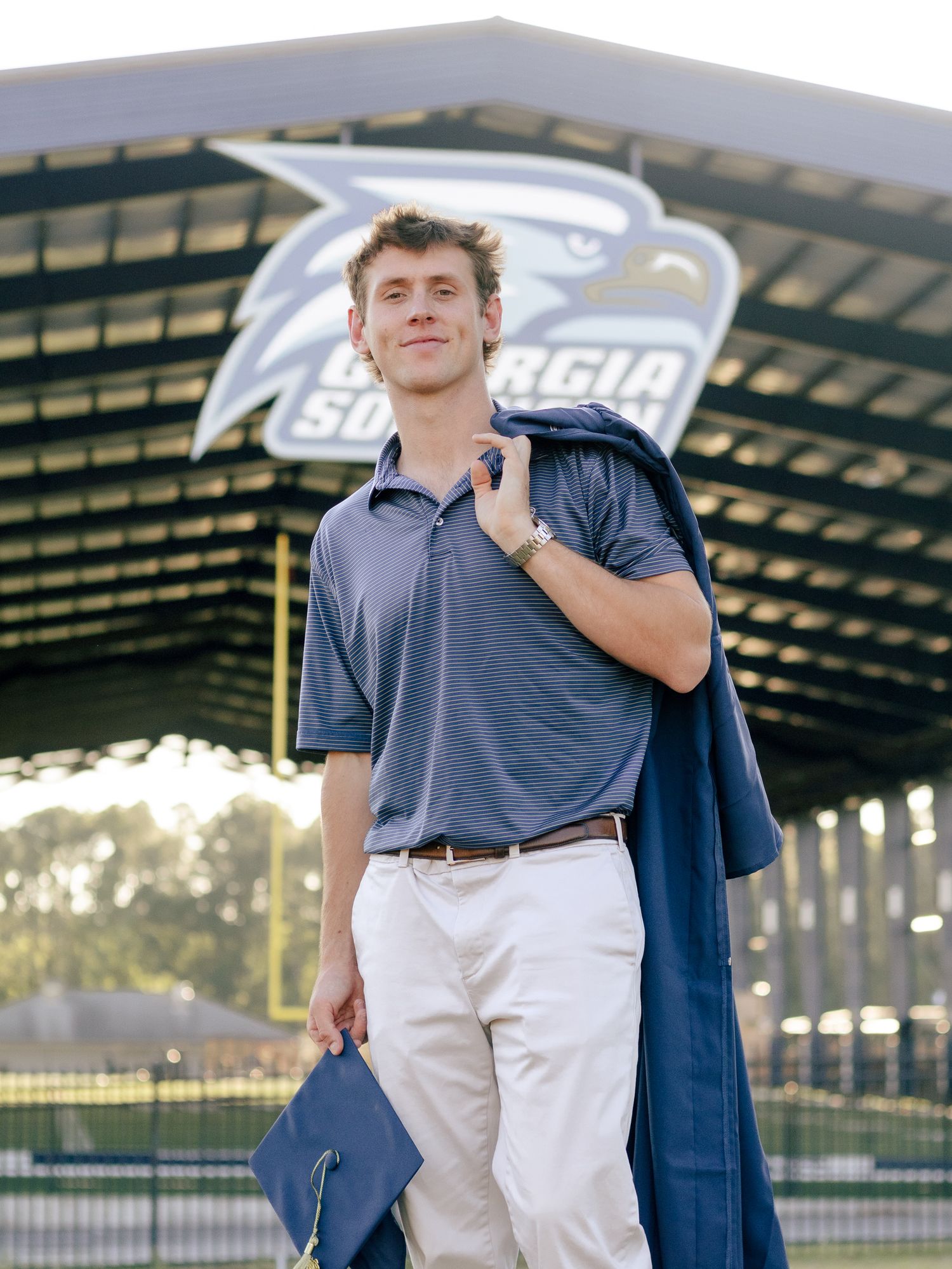 Smiling graduate in a blue polo and khakis holds a cap and gown while standing beneath a Georgia Southern eagle logo on a stadium structure.