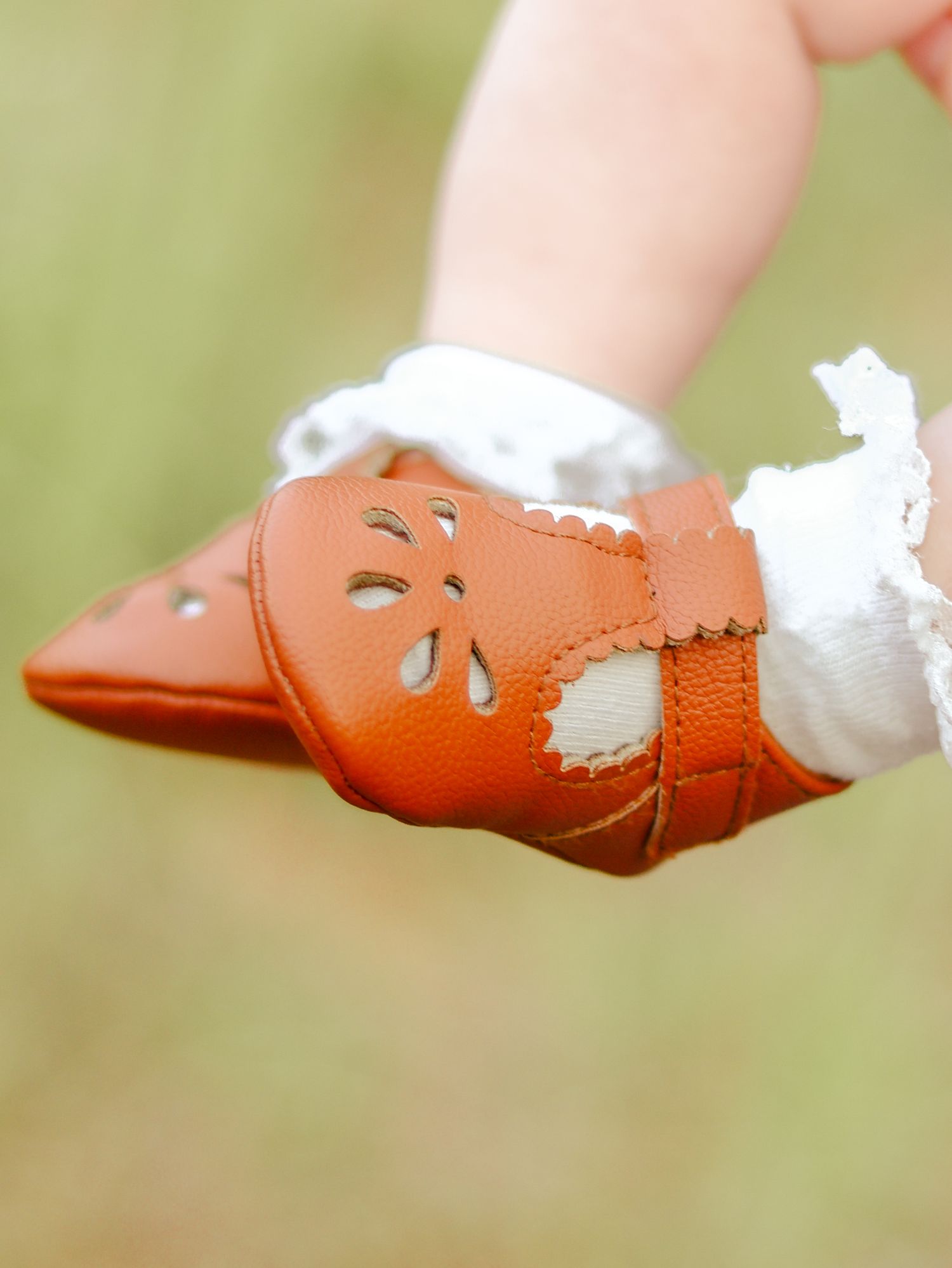 Close-up of a baby's foot in a tan leather Mary Jane shoe with cutout flowers and a white lace ruffle sock on a blurred green background.