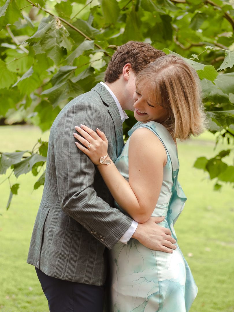 A couple embraces under green tree leaves, the woman in a pale blue dress smiling as she holds the man in a gray plaid blazer.