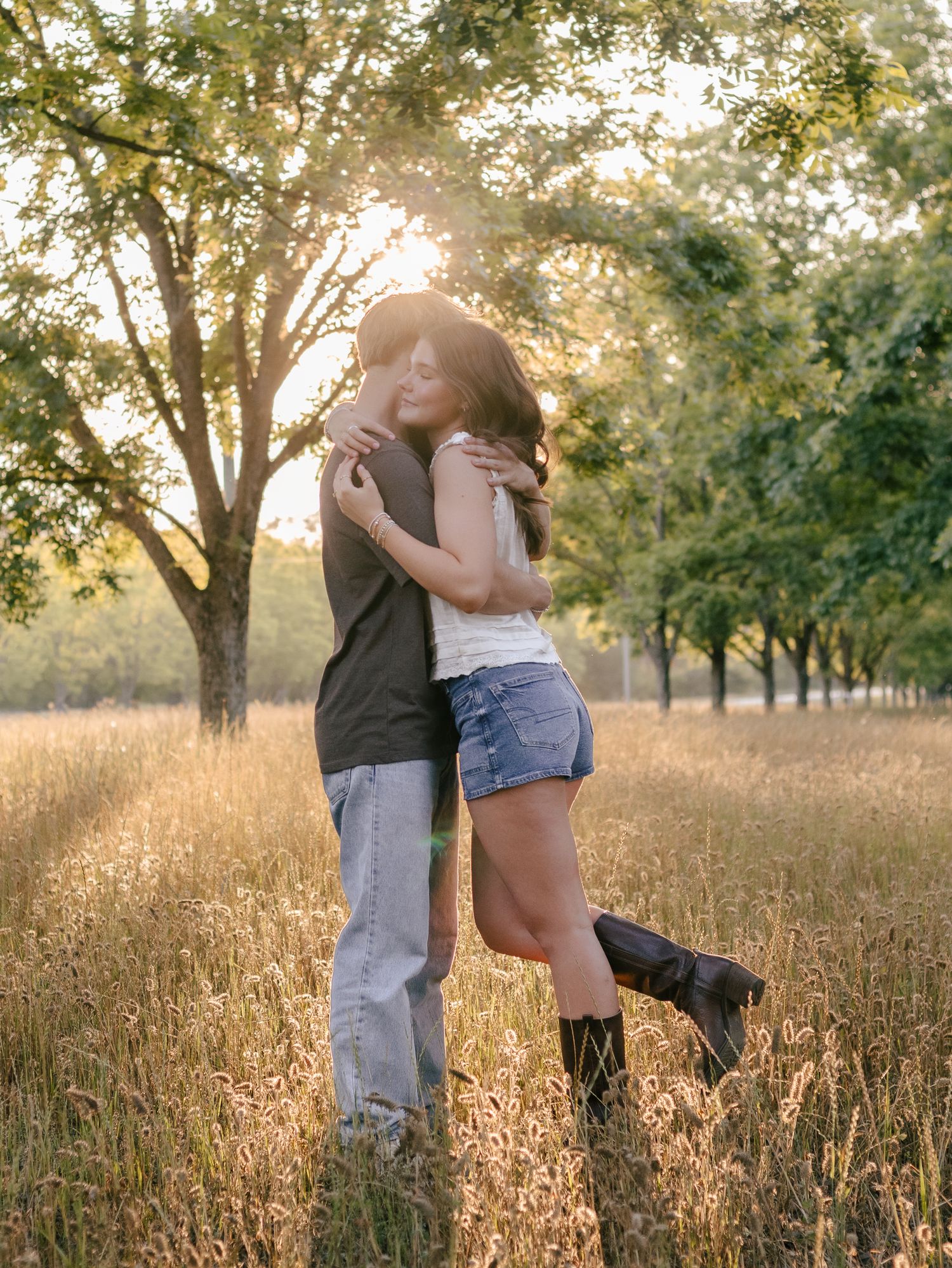 Couple embracing in a golden grass field under tall trees at sunset, the woman in denim shorts and cowboy boots lifting her leg.