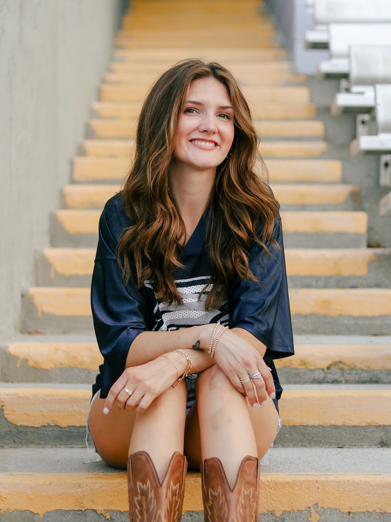 Smiling young woman with long wavy brown hair sits on yellow-edged stadium steps wearing a navy jersey, shorts and tan cowboy boots.