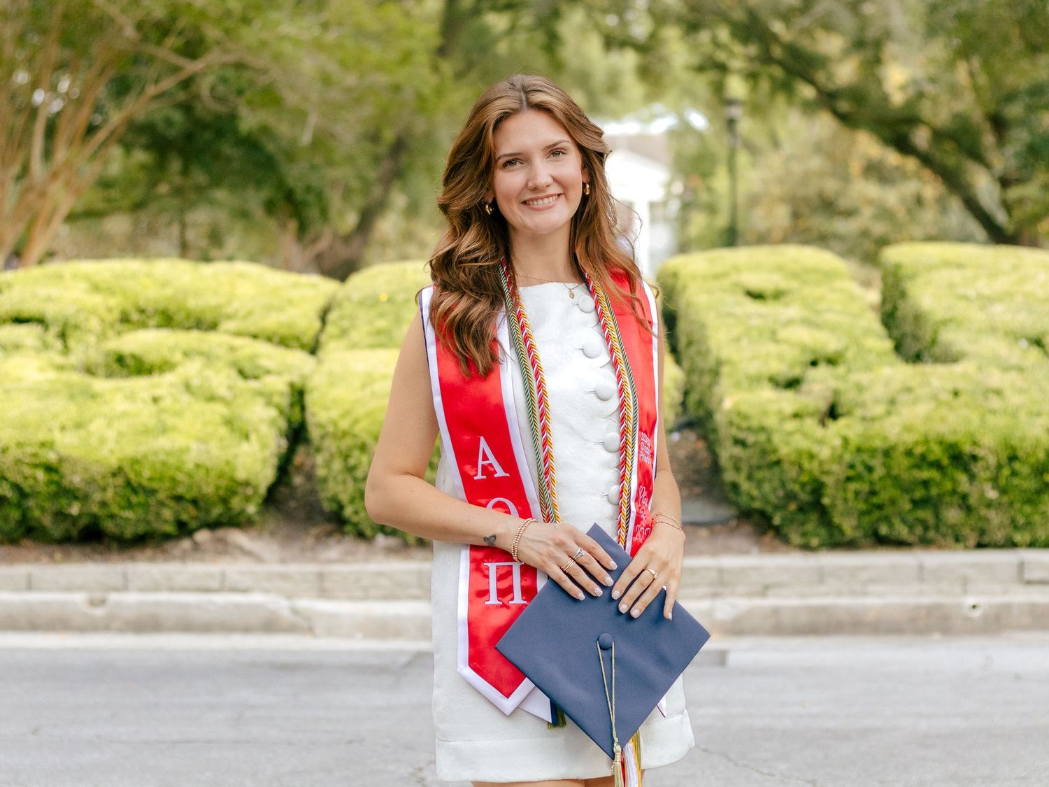 Smiling graduate in a white dress and red stole with Greek letters holds a navy graduation cap while standing near trimmed green hedges.